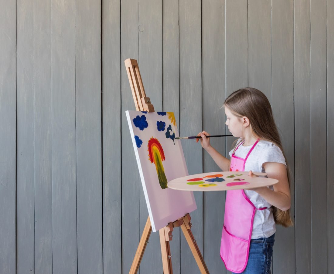 close-up-blonde-girl-painting-with-paint-brush-easel-standing-against-gray-wooden-wall